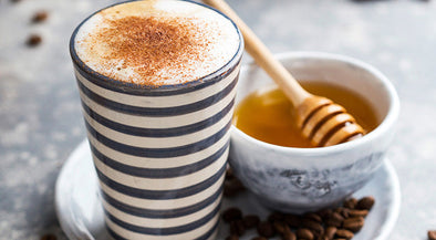 A striped ceramic cup filled with frothy coffee topped with cinnamon, placed next to a small bowl of honey with a wooden honey dipper. Coffee beans are scattered around on a textured surface.